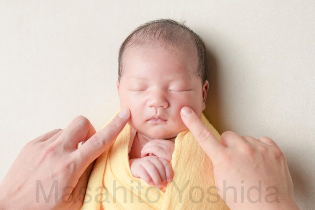 下伊那郡松川町のニューボーンフォト出張撮影｜黄色いおくるみ、白い背景でパパとママにほっぺたをツンツンされている赤ちゃんの写真